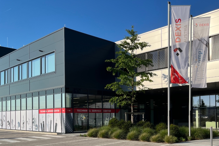 Modernes Industriegebäude mit grauer Fassade. Davor ist eine Grünflächen mit Baum angelegt, wo auch zwei Fahnen stehen mit "DEXIS Austria" und "multiCrimp".  Glaswände geben den Blick auf das Technik und Service Center sowie den Eingangsbereich des Bürogebäudes frei. Es ist strahlend blauer Himmel zu sehen.