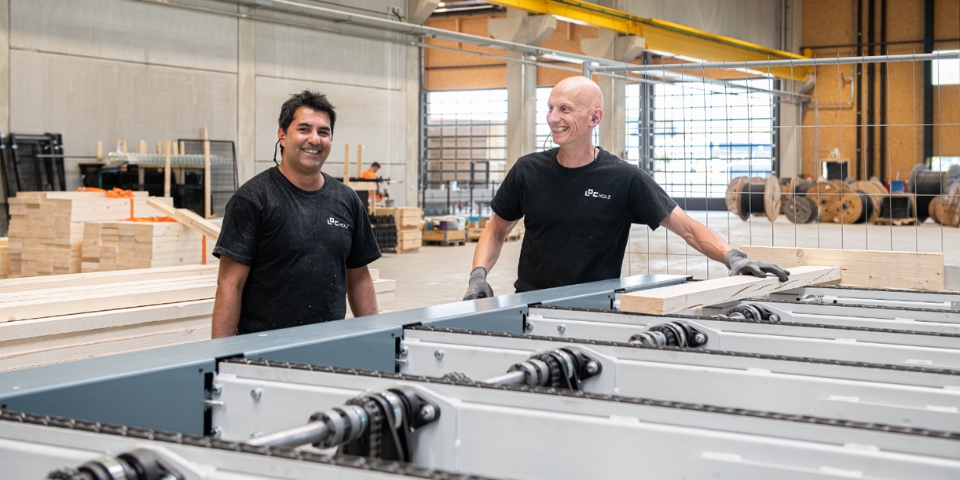 Zwei Arbeiter in schwarzen T-Shirts und Handschuhen lächeln in einer geschäftigen Produktionshalle. Die zwei Männer stehen neben Maschinen, die Holzbretter tragen. Im Hintergrund sieht man Holzstapeln und große Fenster.