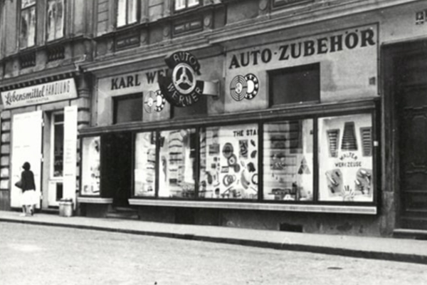 Ein Schwarz-Weiß-Foto zeigt ein historisches Schaufenster mit Autozubehör. Die großen Schaufenster präsentieren die Artikel, unter Schildern und zwei runden Logos.