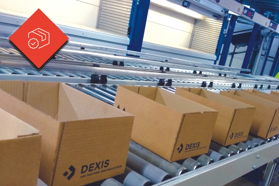 Eine Reihe leerer Kartons mit dem „DEXIS“-Logo sind auf einem Förderband in einer Industrieumgebung aufgereiht. In der oberen linken Ecke ist eine rote Raute mit einem Pakte-Symbol zu sehen.