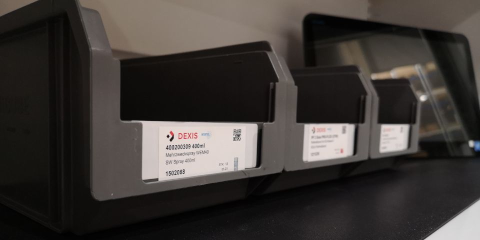 Drei graue Plastikbehälter mit weißen und roten Etiketten mit der Aufschrift „DEXIS“ stehen auf einem RFID-Regal. Die Etiketten geben den Inhalt und Produktdetails an, darunter eine Teilenummer und einen QR-Code. Die Behälter sind ordentlich in einer Reihe angeordnet.