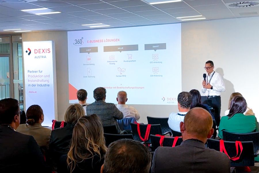 In einem Konferenzraum sitzen Menschen vor einer Leinwand, auf der zwei Moderatoren von DEXIS Austria sprechen. Das aufmerksame Publikum verfolgt die auf die Leinwand projizierten Folien, die das Einsparungspotenziale mit Hilfe von DEXIS präsentieren. Der Raum verfügt über einen Deckenprojektor und weiße Wände.